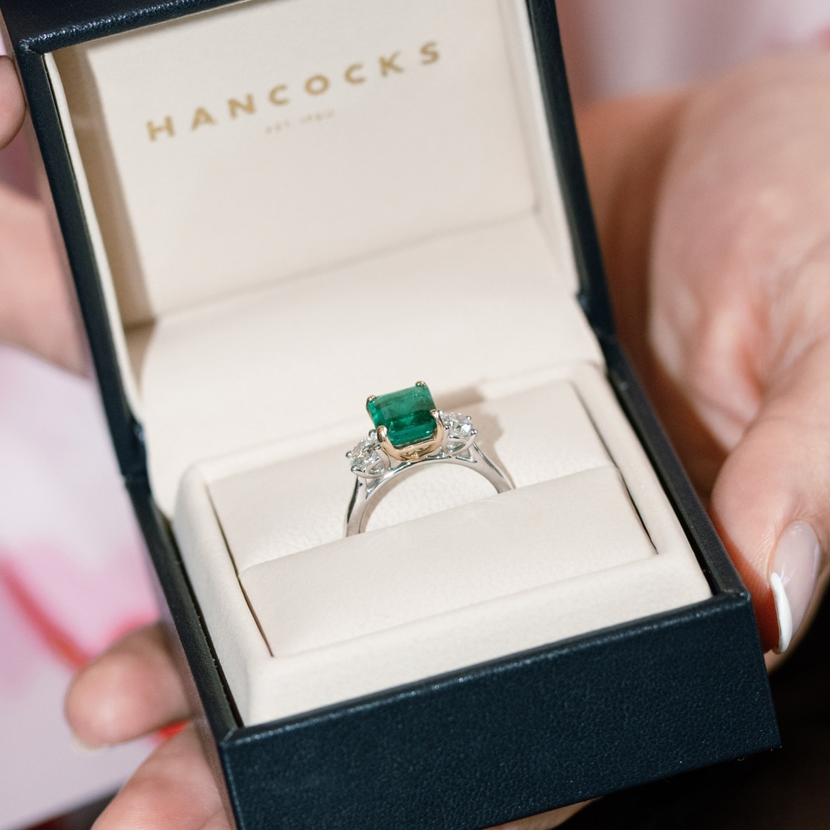 Platinum emerald and diamond ring with an emerald cut emerald and diamond side stones, held in a Hancocks ring box.