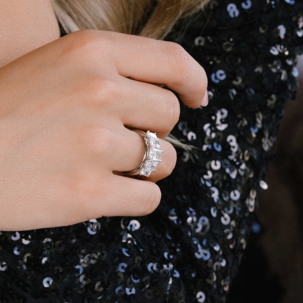 Platinum radiant cut diamond five-stone ring worn on the hand, set against a black sequinned sleeve.