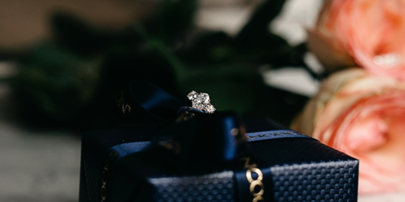 A diamond jewellery ring sitting on top of a black fine jewellery box with two roses behind it. Product photography image.